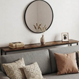 May include: A long, narrow console table with a dark wood-grain surface and a black metal frame, positioned behind a gray sofa. The table holds two stacked books, a light-colored ceramic vase with dried grasses, and a small framed photograph.