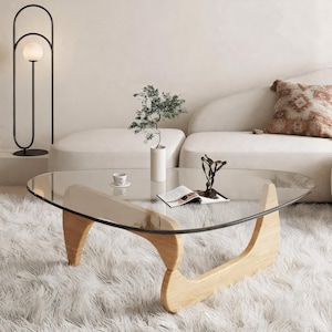 May include: A coffee table featuring a triangular glass top with rounded edges, supported by a light-toned wooden base with curved, sculptural legs. The table is positioned on a textured white rug, holding a small white cup, a book, and a plant.