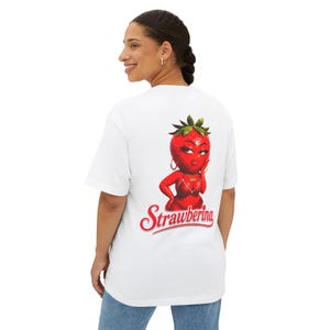 May include: White t-shirt featuring a cartoon strawberry character with red lips, gold hoop earrings, and a red bikini top. The text "Strawbering" is written in red script below the graphic.