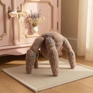 May include: A handmade, crocheted spider toy in shades of brown with teal accents. The plush spider has eight legs and a rounded body. It is sitting on a neutral-colored rug in front of a pink cabinet.