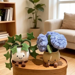 May include: Two plush plant toys with smiling faces sit on a wooden surface. One is a white pot with green ivy, the other is a tan pot with blue flowers. The toys have brown legs and are in a well-lit room.