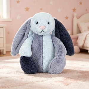May include: A soft toy bunny in blue, grey, and navy hues. The bunny features long, floppy ears, a pink nose, and a fluffy texture. It is positioned on a light pink rug, likely in a child's bedroom.