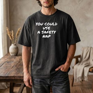 Può includere: T-shirt grigio scuro con la scritta bianca "YOU COULD USE A SAFETY NAP." La maglietta è di vestibilità ampia con maniche corte. La persona indossa jeans grigio scuro e ha la mano in tasca.