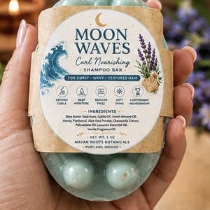 May include: A light blue, oval-shaped shampoo bar with a textured, bumpy surface. It is wrapped in a tan paper label featuring the text Moon Waves Curl Nourishing Shampoo Bar, a wave illustration, lavender sprigs, and a net weight of 5 oz.