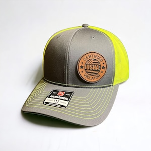 May include: A gray and neon yellow trucker hat with a curved brim. The hat features a circular patch with the text "CERTIFIED OSHA VIOLATOR" and a small American flag design. The brand "Richardson" is visible on a tag.
