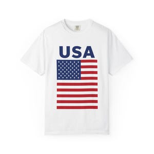 Puede incluir: Camiseta blanca con la palabra "USA" en azul marino sobre un gráfico de la bandera estadounidense. El diseño de la bandera incluye un rectángulo azul con estrellas blancas y rayas rojas y blancas.