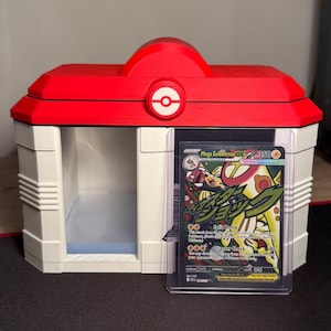 Könnte beinhalten: Ein weiß-rotes 3D-gedrucktes Pokémon-Karten-Display. Das Gehäuse hat ein rotes Dach mit einem weißen Pokéball-Emblem. Eine Pokémon-Karte ist in einer Schutzhülle sichtbar, mit einem Mega-Elektross-Charakter.