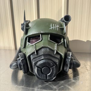 Puede incluir: Un casco táctico de color verde oliva con una máscara respiratoria negra, lentes oscuros para los ojos y un gráfico de marcas de conteo en blanco en la frente. El casco incluye una antena lateral y un tubo cilíndrico en el lado izquierdo.