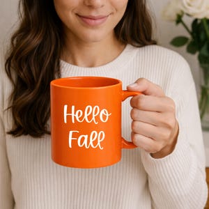 Op de afbeelding: Een oranje keramische mok met de woorden "Hello Fall" in witte letters. De mok heeft een oor en wordt vastgehouden door een persoon die een witte trui draagt. De mok is een seizoensartikel.