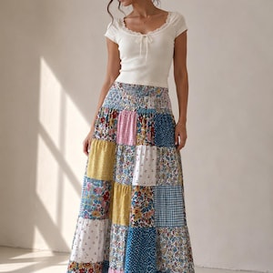 Peut inclure: Un haut blanc côtelé à manches courtes avec une encolure dégagée bordée de dentelle et un détail à nouer, associé à une jupe longue en patchwork composée de divers carrés de tissu à motifs floraux, vichy et unis dans des tons bleu, jaune, rose et blanc.