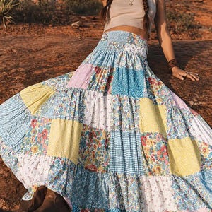 Puede incluir: Falda larga de patchwork con una variedad de estampados florales, cuadros y lunares en tonos azules, amarillos, rosas y blancos. La falda tiene una cintura fruncida y un diseño fluido y escalonado. Se ven botas marrones en la parte inferior de la imagen.