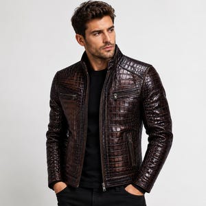 Puede incluir: Una chaqueta de cuero marrón oscuro con un patrón texturizado de piel de cocodrilo, cuello alto y cremallera frontal completa. La chaqueta incluye dos bolsillos horizontales con cremallera en el pecho y dos bolsillos laterales inferiores con herrajes metálicos a juego.