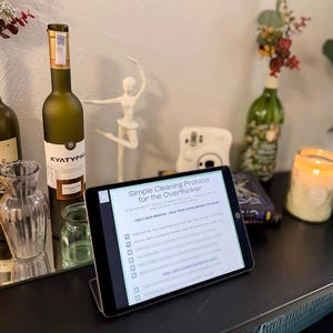 May include: A tablet displaying a "Simple Cleaning Protocol for the Overthinker" sits on a stand. The image includes a wine bottle, a ballerina figurine, a candle, and other decorative items, suggesting a lifestyle or home decor theme.