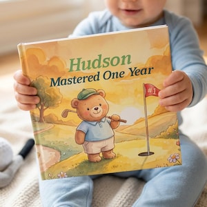 May include: A hardcover book titled Hudson Mastered One Year featuring a watercolor illustration of a brown bear wearing a green cap and blue shirt on a golf course with a red flag marked number one, set against a soft yellow sunset sky.