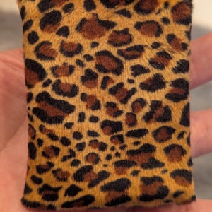Puede incluir: Un objeto rectangular de felpa con estampado de leopardo. La tela es una mezcla de beige, marrón y negro, con manchas de diferentes tamaños y formas. El objeto tiene una textura suave y peluda, y los bordes están bien acabados.