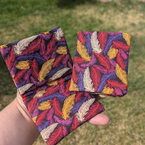 May include: Two square fabric pouches featuring a repeating pattern of feathers in vibrant shades of red, purple, yellow, and cream against a black background. The pouches are constructed from a soft textile material with visible stitching along the edges.