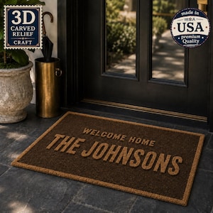 May include: A rectangular brown coir doormat featuring the text WELCOME HOME THE JOHNSONS in raised, carved relief lettering. The mat is positioned on a dark stone floor in front of a black door, with a brass umbrella stand and a potted plant nearby.