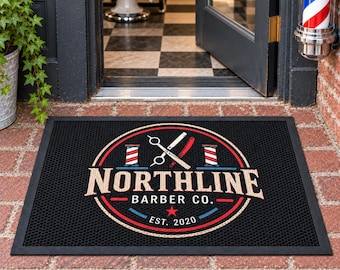 May include: A rectangular black doormat featuring a circular logo with red, white, and blue accents. The design includes crossed barber shears, two barber poles, and the text NORTHLINE BARBER CO. EST. 2020 with a small red star centered below.