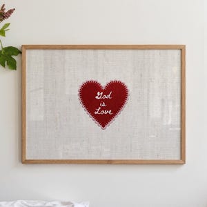 Puede incluir: Una pieza de arte mural enmarcada que presenta un corazón de fieltro rojo centrado sobre un fondo de tela texturizada de color beige claro. El corazón tiene un borde festoneado cosido en rosa y el texto bordado en blanco God is Love.