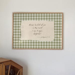 Op de afbeelding: Een rechthoekig ingelijst wandkunstwerk met een lichtgroen en wit ruitjespatroon op de achtergrond. In het midden bevindt zich een gebroken wit papier met gescheurde randen en de zwarte handgeschreven tekst: Always be full of joy in the Lord I say it again REJOICE! — philippians 4:4.