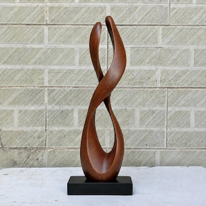 Peut inclure: Une grande sculpture en bois brun lisse présentant une forme abstraite, torsadée et fluide qui s'incurve vers le haut jusqu'à une pointe. La sculpture est montée sur une base noire rectangulaire solide, créant une silhouette verticale.