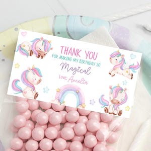 May include: A rectangular white favour tag featuring pastel-coloured illustrations of unicorns, a rainbow, and stars. The text reads: THANK YOU FOR MAKING MY BIRTHDAY SO Magical Love, Amelia. The tag is attached to a clear plastic bag filled with pink sweets.