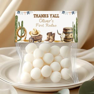 May include: A clear plastic treat bag filled with round white candies, topped with a cardboard header featuring the text THANKS YALL Oliver's First Rodeo, illustrated with a lasso, cowboy hat, wagon, boots, bull skull, barrel, and cacti.