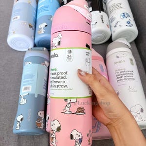 K&ouml;nnte beinhalten: Eine Auswahl an Owala-Isolierwasserflaschen in verschiedenen Farben, darunter Rosa, Blau und Wei&szlig;. Jede Flasche ist mit einem Snoopy-Cartoon-Design versehen. Die Flaschen haben einen eingebauten Strohhalm und sind auslaufsicher. Der Text lautet: "auslaufsicher, isoliert und mit eingebautem Strohhalm."