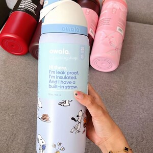 K&ouml;nnte beinhalten: Eine Sammlung von Owala-Wasserflaschen in verschiedenen Farben, darunter Rot, Rosa und Blau. Eine Flasche zeigt ein Snoopy-Cartoon-Design und den Text "Hi there. I'm leak proof. I'm insulated. And I have a built-in straw."