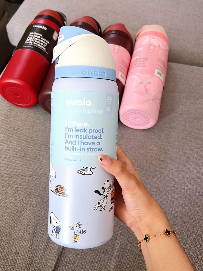K&ouml;nnte beinhalten: Eine Sammlung von Owala-Wasserflaschen in verschiedenen Farben, darunter Rot, Rosa und Blau. Eine Flasche zeigt ein Snoopy-Cartoon-Design und den Text "Hi there. I'm leak proof. I'm insulated. And I have a built-in straw."