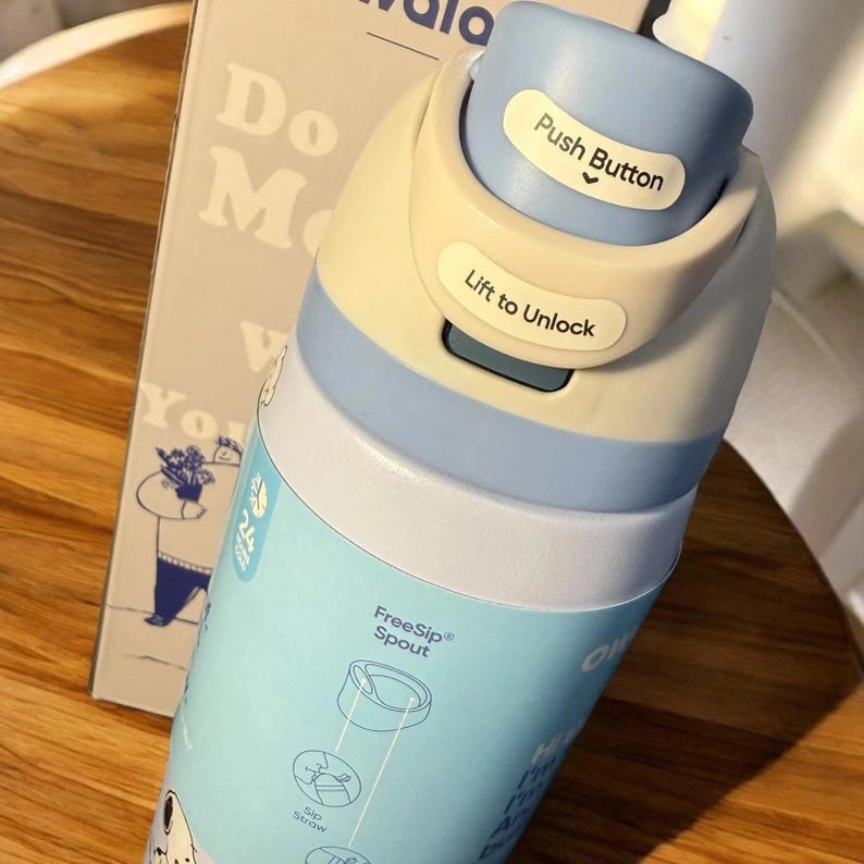 K&ouml;nnte beinhalten: Eine hellblaue und cremefarbene Wasserflasche mit einem Druckknopfdeckel und den W&ouml;rtern "Push Button" und "Lift to Unlock" darauf. Die Flasche hat ein Cartoon-Design und die Worte "FreeSip Spout" sind sichtbar.
