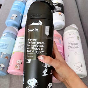 K&ouml;nnte beinhalten: Schwarze Owala-Wasserflasche mit Snoopy-Illustrationen und Text. Die Flasche hat 945 ml, ist auslaufsicher, isoliert und hat einen eingebauten Strohhalm. Weitere Flaschen mit Snoopy-Designs sind im Hintergrund zu sehen.