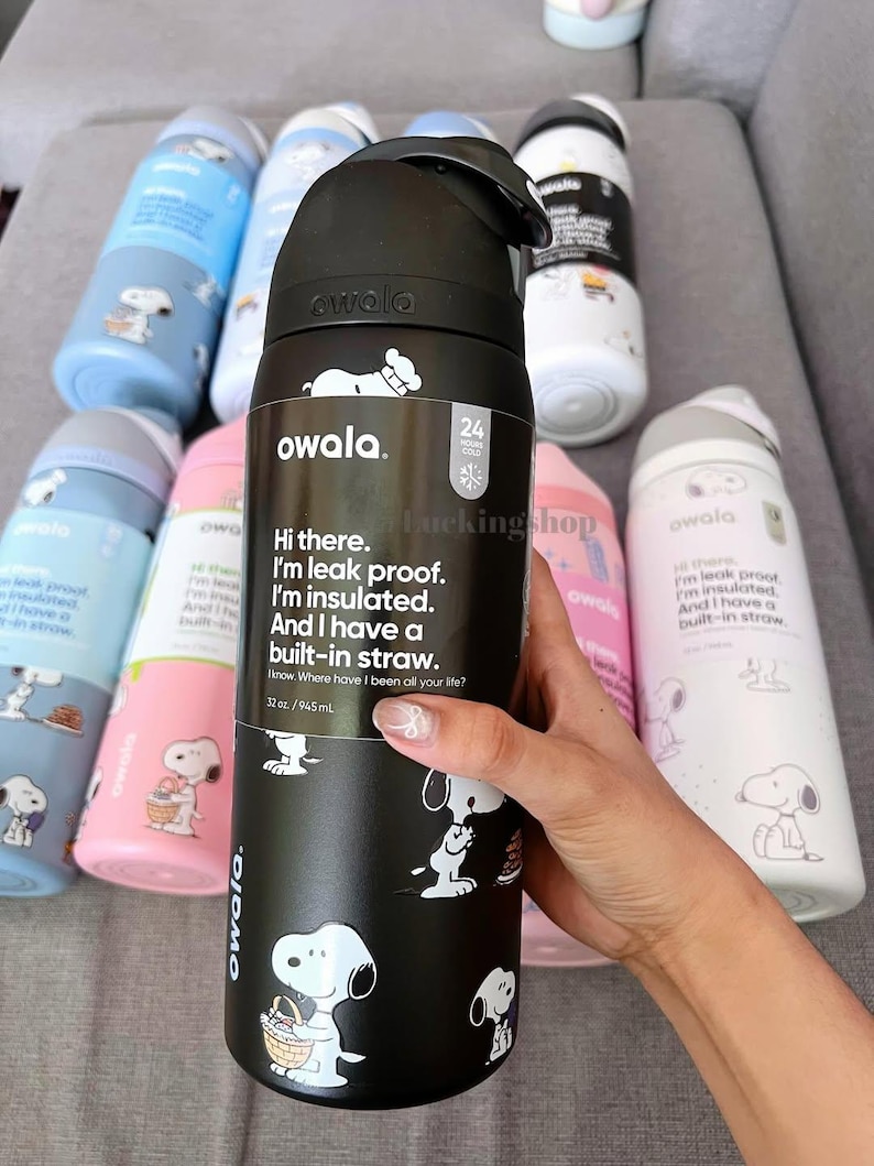 K&ouml;nnte beinhalten: Schwarze Owala-Wasserflasche mit Snoopy-Illustrationen und Text. Die Flasche hat 945 ml, ist auslaufsicher, isoliert und hat einen eingebauten Strohhalm. Weitere Flaschen mit Snoopy-Designs sind im Hintergrund zu sehen.