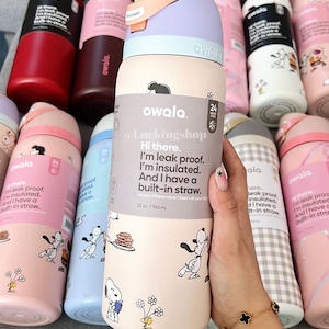 K&ouml;nnte beinhalten: Eine Sammlung von Owala-Isolierwasserflaschen in verschiedenen Farben, darunter Rot, Rosa und Blau. Die Flaschen zeigen ein Snoopy-Cartoon-Design und den Text: "I'm leak proof. I'm insulated. And I have a built-in straw."