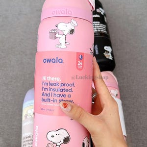 K&ouml;nnte beinhalten: Rosa Owala-Wasserflasche mit Snoopy-Illustrationen. Die Flasche ist isoliert und hat einen eingebauten Strohhalm. Der Text auf der Flasche lautet: "Hi there. I'm leak proof. I'm insulated. And I have a built-in straw." Sie fasst 945 ml.