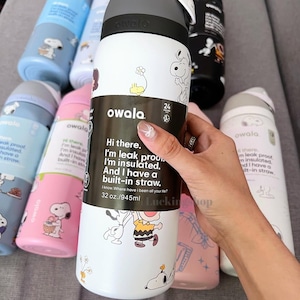 K&ouml;nnte beinhalten: Wei&szlig;e Owala-Wasserflasche mit schwarzem und grauem Deckel, mit Snoopy- und Charlie Brown-Illustrationen. Die Flasche hat einen eingebauten Strohhalm und fasst 945 ml. Weitere Flaschen in Blau, Rosa und Schwarz sind sichtbar.