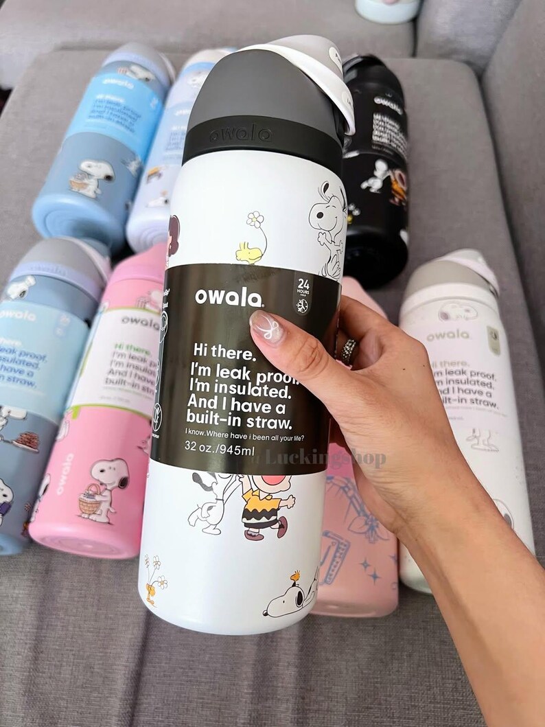 K&ouml;nnte beinhalten: Wei&szlig;e Owala-Wasserflasche mit schwarzem und grauem Deckel, mit Snoopy- und Charlie Brown-Illustrationen. Die Flasche hat einen eingebauten Strohhalm und fasst 945 ml. Weitere Flaschen in Blau, Rosa und Schwarz sind sichtbar.