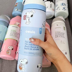 K&ouml;nnte beinhalten: Owala-Wasserflaschen in verschiedenen Farben mit Snoopy-Designs. Die Flaschen sind hellblau, rosa und wei&szlig;, mit Snoopy-Illustrationen und Text. Sie haben einen eingebauten Strohhalm und ein Fassungsverm&ouml;gen von 945 ml.