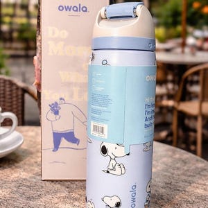 K&ouml;nnte beinhalten: Eine hellblaue Owala-Trinkflasche mit wei&szlig;em und hellblauem Deckel, mit Snoopy-Illustrationen. Die Flasche hat ein Fassungsverm&ouml;gen von 385 ml und wird mit einer passenden Box geliefert. Auf der Box stehen das Owala-Logo und die Worte "Do More What You Love."
