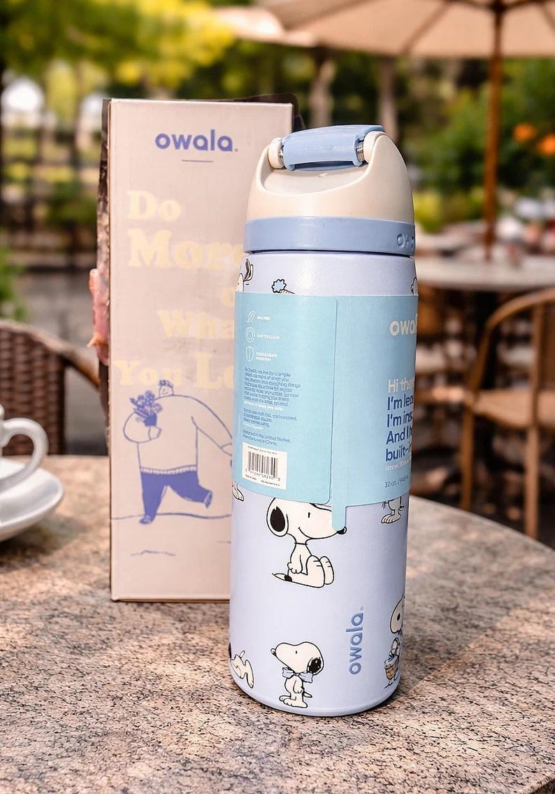 K&ouml;nnte beinhalten: Eine hellblaue Owala-Trinkflasche mit wei&szlig;em und hellblauem Deckel, mit Snoopy-Illustrationen. Die Flasche hat ein Fassungsverm&ouml;gen von 385 ml und wird mit einer passenden Box geliefert. Auf der Box stehen das Owala-Logo und die Worte "Do More What You Love."