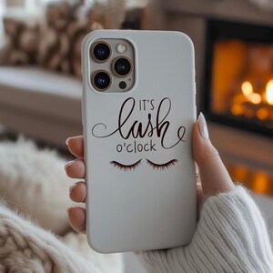 Może przedstawiać: Jasnoszara obudowa telefonu z napisem "It's lash o'clock" w ozdobnej czcionce oraz parą zamkniętych rzęs. Obudowa posiada moduł aparatu z trzema obiektywami.