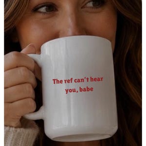 May include: A white ceramic mug featuring the red text "The ref can't hear you, babe" printed on the front. The mug has a smooth, cylindrical body with a curved handle on the side, designed for holding hot beverages in a standard, functional style.