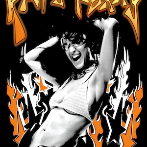 May include: A black graphic t-shirt featuring a high-contrast, halftone-style image of a person with hands behind their head. The design includes orange and yellow flames, black star shapes, and the text KATY PERRY in a stylized orange font.