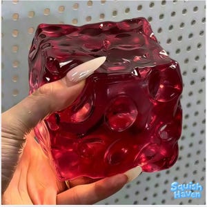 May include: A translucent, deep red cube with a textured, dimpled surface featuring circular indentations on each face. The object has a glossy, reflective finish and a squishy appearance, with the text Squish Haven visible in the bottom corner.