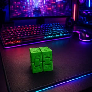 May include: A vibrant green fidget cube rests on a black desk mat with a glowing blue and purple edge. A backlit keyboard and mouse are in the background, with a cityscape on the monitor. The cube is a two-piece design.