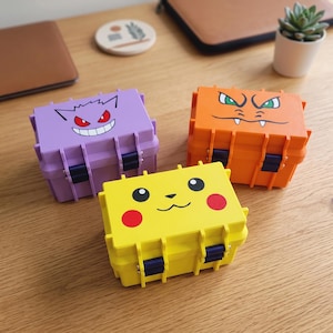 May include: Three rectangular storage cases with latch closures, each featuring a different character face: a purple case with a Gengar design, an orange case with a Charizard design, and a yellow case with a Pikachu design, all with dark blue latches.
