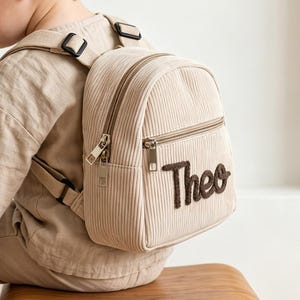May include: A beige corduroy mini backpack featuring a front zippered pocket with the name Theo embroidered in dark brown script. The backpack has adjustable shoulder straps and silver-toned metal zippers, worn by a child in beige clothing.