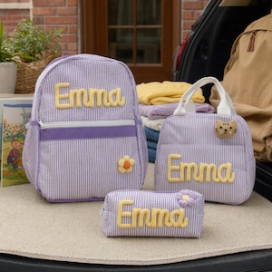 May include: A purple and white striped backpack, lunch bag, and pencil case set. Each item features the name Emma in yellow, textured, raised lettering. The backpack has a small flower patch, while the lunch bag includes a cat face patch.