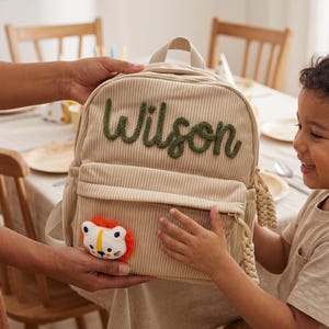 May include: A beige corduroy backpack featuring the name Wilson embroidered in dark green yarn across the top. The front pocket is adorned with a small, plush lion face patch with an orange mane. The zipper pull has a braided cord detail.
