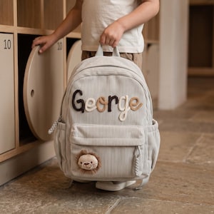 May include: A light gray corduroy backpack featuring the name George embroidered in brown and beige yarn across the front. The bag includes a front zippered pocket adorned with a small, plush lion face patch and matching braided zipper pulls.